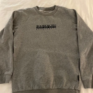 Napapijri tröja  - Säljer en stilren grå sweatshirt från Napapijri med deras logga tryckt på bröstet. Tröjan har långa ärmar och ribbade muddar vid ärmslut och nederkant. Perfekt för en avslappnad look. Storlek S