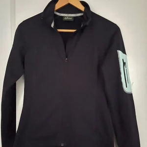 Svart långärmad tröja från Peak Performance - Snygg svart långärmad tröja, softcheln,  från Peak Performance med en ljusblå detalj på ärmen. Perfekt för en sportig look. Tröjan har en dragkedja vid halsen och är tillverkad i ett syntetmaterial som ger en skön passform. 