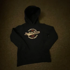 Blå hoodie från Jack & Jones - Säljer en blå hoodie från Jack & Jones med tryck i vitt och gult på bröstet. Tröjan har en klassisk design med huva och långa ärmar. Perfekt för en avslappnad stil.