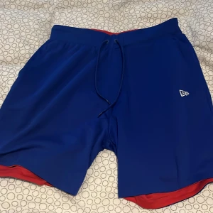 Vändbara shorts från New Era - Snygga vändbara shorts från New Era i blått och rött. Perfekta för en sportig look med elastisk midja och snörning för bästa passform. Materialet är lätt och bekvämt. Köpa 2020 o har inte blivit använda på långt tag. Pris går att diskutera. Original pris runt 500