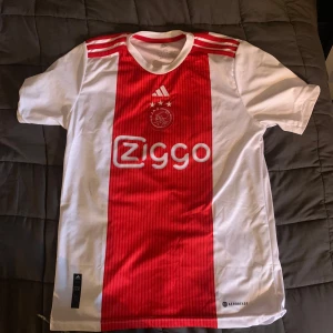 Vit och röd fotbollströja från Adidas - Säljer en snygg vit och röd fotbollströja från Adidas med Ajax-logga och Ziggo-sponsortryck. Tröjan har korta ärmar och ett klassiskt randigt mönster i rött på framsidan. Perfekt för fotbollsfans som vill visa sitt stöd för laget storleken på tröjan är S, 178 men skulle säga att den passar dem som är 170 också 