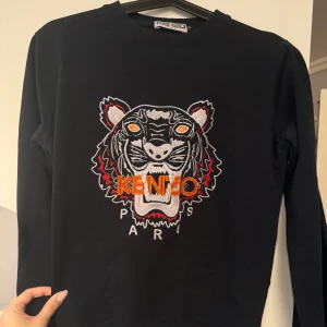 Svart sweatshirt från Kenzo med tigerbroderi - Säljer en svart sweatshirt från Kenzo som är AKopia med ett tigerbroderi i flera färger på framsidan. Tröjan har långa ärmar och en rund halsringning. Perfekt för en stilren och trendig look.