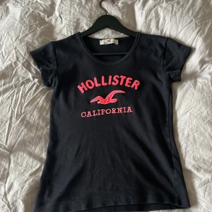 Svart t-shirt från Hollister - Snygg svart t-shirt från Hollister med råsa text och logga på bröstet. Perfekt för en avslappnad stil med korta ärmar och rund halsringning.