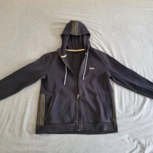 Boss hoodie - Snygg mörkblå huvtröja från Boss med dragkedja och randiga detaljer på fickorna och huvan. Tröjan har långa ärmar och en bekväm passform. Perfekt för en avslappnad stil. 