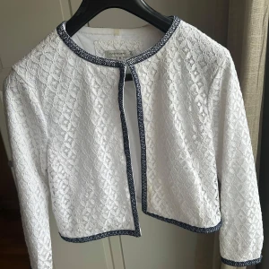 Vit spetskofta - Säljer en elegant vit spetskofta med en fin spetsdesign med mörkblå kantdetaljer och den är långärmad. Perfekt till våren!
