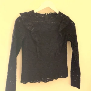 Svart spetsblus från Zara - Elegant svart spetsblus från Zara med långa ärmar och volangdetaljer vid axlarna. Perfekt för en stilren look. 