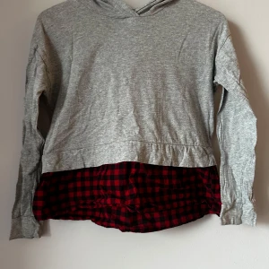 Grå hoodie med röd- och svartrutig detalj - Säljer en grå hoodie med en cool röd- och svartrutig detalj längst ner. Tröjan har långa ärmar och en bekväm huva. Perfekt för en avslappnad stil. Står storlek M men skulle säga 130-140.