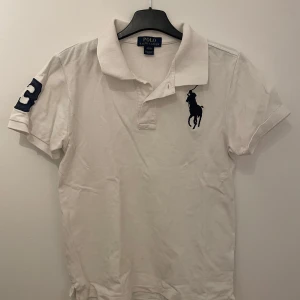 Vit pikétröja från Ralph Lauren - Säljer en klassisk vit pikétröja från Ralph Lauren med korta ärmar. Tröjan har en broderad logga på bröstet och en stor siffra på ärmen. Perfekt för en stilren och sportig look. Skriv om ni har funderingar och priset är inte hugget i sten!