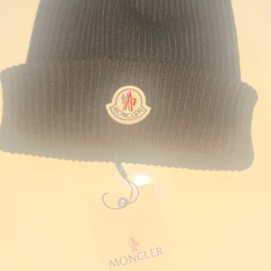 Svart ribbad mössa från Moncler - Snygg svart ribbad mössa från Moncler med broderad logotyp framtill. Perfekt för att hålla värmen under kyliga dagar. Klassisk design som passar till det mesta.