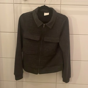 Svart tröja från Vila - Snygg svart tröja från Vila med dragkedja och krage. Tröjan har långa ärmar och två framfickor med lock. Perfekt för en stilren look.