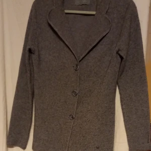 Grå kappa med hjärtdetalj - Säljer en stilren grå kappa med knappar och en unik hjärtdetalj. Jackan är tillverkad i en blandning av viskos, polyester och nylon, vilket ger en mjuk och bekväm känsla. Perfekt för kyligare dagar med sin långa ärm och klassiska design.