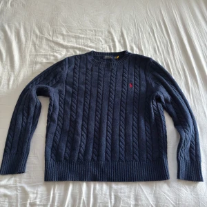 Mörkblå kabelstickad tröja från Ralph Lauren - Säljer en stilren mörkblå kabelstickad tröja från Ralph Lauren. Tröjan har en klassisk rund halsringning och ett diskret rött logotypbroderi på bröstet. Perfekt för kyligare dagar. Storlek XL men passar som M.