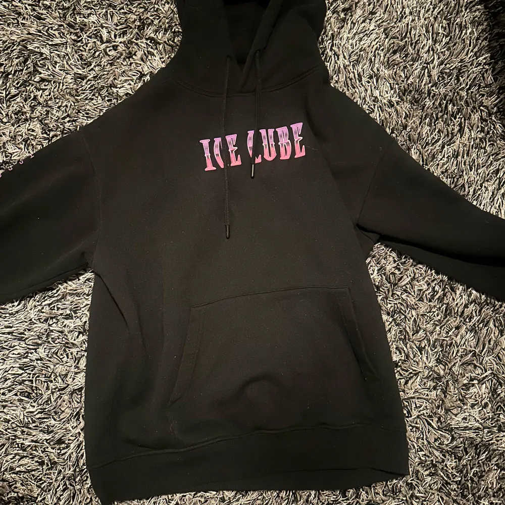 Cool svart hoodie med rosa text 'ICE CUBE' på framsidan och 'TODAY WAS A GOOD DAY' längs ärmarna. Baksidan har ett stort tryck av Ice Cube i grått och rosa. Perfekt för fans av hiphop och streetwear.✨💫💫. Neuletakit & Villapaidat.