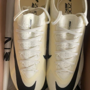 Nike Zoom Mercurial i vitt och svart - Säljer ett par Nike Zoom Mercurial i vitt med svarta detaljer. Skorna har en strömlinjeformad design med snörning och är perfekta för sportiga aktiviteter. De är tillverkade i syntetmaterial för en lätt och bekväm känsla.