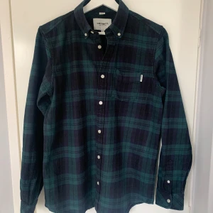 Grön rutig skjorta från Carhartt - Snygg grön och blå rutig skjorta från Carhartt med klassisk krage och knappar framtill. Skjortan har en bröstficka med en liten etikett. Perfekt för en avslappnad stil.