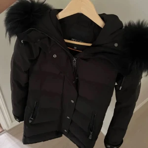 Svart dunjacka från Hollies - Säljer en snygg svart dunjacka från Hollies med päls huva. Jackan har dragkedja och knappar framtill samt praktiska fickor med dragkedja. Perfekt för kalla dagar och varm. Jackan är i perfekt skick! 