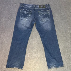 True Religion Jeans - Snygga blå jeans från True Religion med unika broderade bakfickor. De har en klassisk femficksdesign och en bekväm passform. Perfekta för en avslappnad stil.