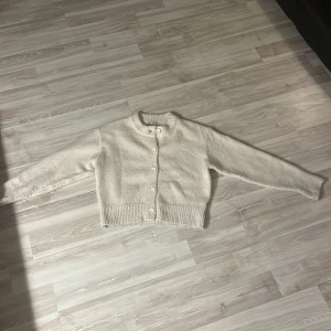 Vit stickad kofta med knappar - Säljer en mysig vit stickad kofta med knappar framtill. Koftan har långa ärmar och ribbade muddar vid ärmslut och nederkant. Perfekt för kyliga dagar!