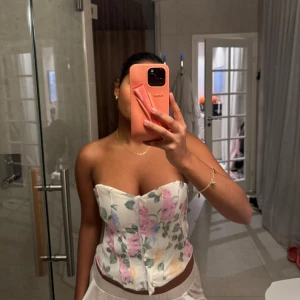 Blommig korsettopp - Säljer en söt blommig korsettopp med pastellfärgade blommor i rosa, lila och gult på en vit bakgrund. Toppen är axelbandslös och har en figurnära passform. Perfekt för en somrig look!