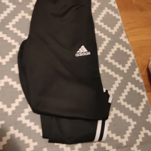 Svarta träningsbyxor från Adidas - Säljer ett par svarta träningsbyxor från Adidas med de klassiska tre vita ränderna längs sidorna. Byxorna har en elastisk midja och är perfekta för sport och träning. Adidas-loggan är broderad på framsidan.