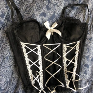Svart korsett med snörning och spetsdetaljer - Elegant svart korsett med vita snörningar och spetsdetaljer. Den har justerbara axelband. OANVÄND