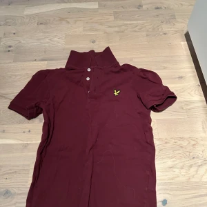 Vinröd pikétröja från Lyle & Scott - Säljer en snygg vinröd pikétröja från Lyle & Scott med klassisk krage och knappar framtill. Tröjan har en broderad logga på bröstet och korta ärmar. Perfekt för en stilren look.