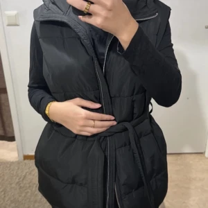 Svart dunväst från Nelly - Säljer en snygg svart dunväst från Nelly. Den har en dragkedja framtill och ett bälte i midjan för en mer figurnära passform. Perfekt för höst och vinter, och den är i nyskick med etiketten kvar. Västen har en huva som ger extra värme och stil.