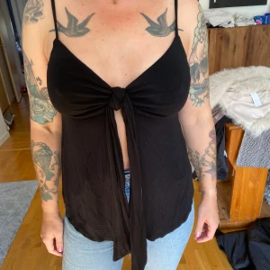 Svart ärmlös topp med knyt - Säljer en snygg svart ärmlös topp med smala axelband och knyt framtill. Toppen har en avslappnad passform och är perfekt för sommarens varma dagar. Den är gjord i ett mjukt och bekvämt material.