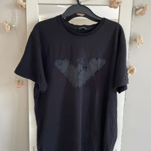 Svart t-shirt från Emporio Armani - Säljer en snygg svart t-shirt från Emporio Armani. Den har en cool logga på framsidan och är i storlek XXL. Perfekt för en stilren och avslappnad look. Materialet känns mjukt och skönt mot huden..personen har medium .har kostat 2000kr
