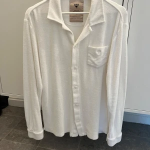 Vit frottéskjorta från OAS - Säljer en supermjuk och bekväm vit skjorta från OAS, perfekt att dra på sig efter ett dopp. Den är gjord av 100% frotté bomull och har en klassisk krage med knappar hela vägen ner. Skjortan har en bröstficka och långa ärmar, vilket gör den både stilren och praktisk. Designad i Sverige och tillverkad i Portugal. Perfekt för sommarens alla äventyr! 🌊