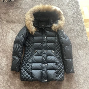 Fin vinterjacka från Rockandblue 🌟 - Figursydd modell i äkta dun Quiltat yttertyg i vattenavvisande material Avtagbar luva med avtagbar fuskpäls Försluts med dragkedja Två framfickor med dragkedja, liten ficka på vänster ärm Bröstficka med dragkedja Två innerfickor med dragkedja 