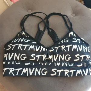 Svart sporttopp med textmönster - Säljer en ny svart sporttopp med vitt textmönster. Från bershka storlek S. Toppen har smala axelband och en bekväm passform, perfekt för träning eller en aktiv vardag. Den är i bra skick och redo för nya äventyr!