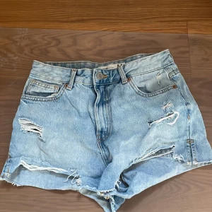 Blå jeansshorts med slitningar - Säljer ett par snygga blå jeansshorts från F2 i storlek M. De har en cool, sliten look med flera revor och fransiga kanter. Perfekta för en avslappnad sommarstil!