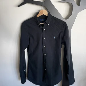 Ralph Lauren Skjorta - Trendig och stilren svart skjorta från Polo Ralph Lauren. Storlek slim fit S. Den svarta färgen är lite ljusare än från fabrik men helhetsintrycket är fortfarande väldigt bra. Skick 8,5/10 nypris 1795kr. Modellen är 183cm och 80kg (sitter rätt tight)
