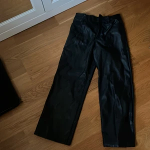 Svarta skinnbyxor - Säljer ett par snygga svarta skinnbyxor i högmidjad modell. De har en dragkedja och knapp framtill samt fickor bak. Perfekta för en cool och stilren look!