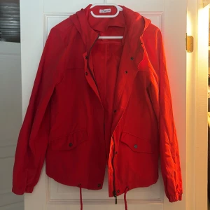 Röd vindjacka  - Säljer en snygg röd vindjacka. Den har en dragkedja framtill och praktiska fickor med knappar. Jackan är nästintill oanvänd och har även luva.