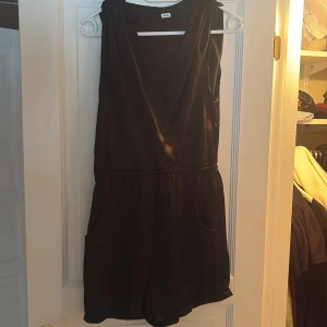 Svart byxdress - Snygg svart ärmlös byxdress i ett mjukt och glansigt material. Den har en djup v-ringning och är i storlek L
