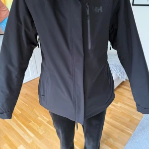 Svart jacka från Helly Hansen - Säljer en svart jacka från Helly Hansen i mycket bra skick. Den har en dragkedja framtill och en hög krage för extra skydd mot väder och vind. Perfekt för höst- och vinterdagar. Jackan är stilren och passar till det mesta. 🖤