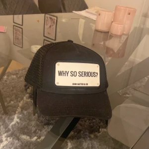 Svart keps från John Hatter & Co - Säljer en cool svart keps från John Hatter & Co med texten 'WHY SO SERIOUS?' på framsidan. Kepsen har ett nätmaterial baktill för bra ventilation och en justerbar rem för perfekt passform. Perfekt för en avslappnad stil eller som statement piece.