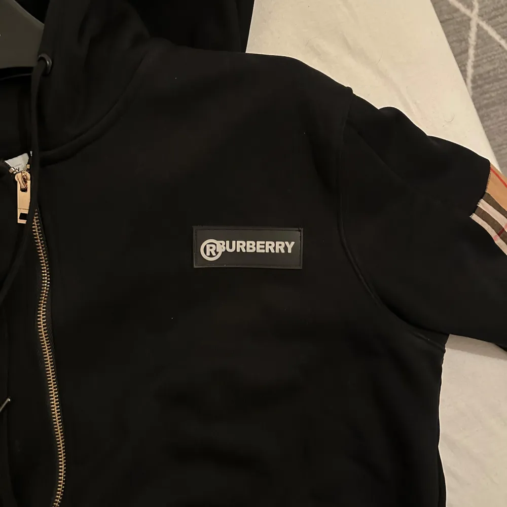 Säljer en snygg svart hoodie från Burberry med dragkedja och klassiska rutiga detaljer på ärmarna. Perfekt för höst och vintern! Den har en bekväm passform och är i mycket bra skick. Perfekt för dig som vill ha en stilren och lyxig look.. Tröjor & Koftor.