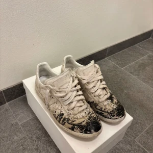 Maison Margiela - Unika Maison Margiela sneakers i vitt med coola svarta färgstänk. Skorna har en klassisk snörning och är i bra skick trots den avsiktliga slitna looken. Perfekta för dig som vill sticka ut med en grisch stil! Buda gärna och tar även emot byten!!