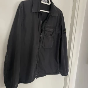 Svart jacka från Stone Island - Säljer en svart jacka från Stone Island i bra skick. Den har en dragkedja framtill och en bröstficka med knapp. Perfekt för höst och vår med sin stilrena design och bekväma passform. Jackan har också det klassiska Stone Island-märket på ärmen. Skick 8/10