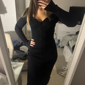 Svart ribbad klänning - Säljer en snygg svart ribbad klänning med lång ärm och knappar framtill. Den har en v-ringad design och är perfekt för både vardag och fest. Klänningen är i bra skick och passar till höst och vinter.