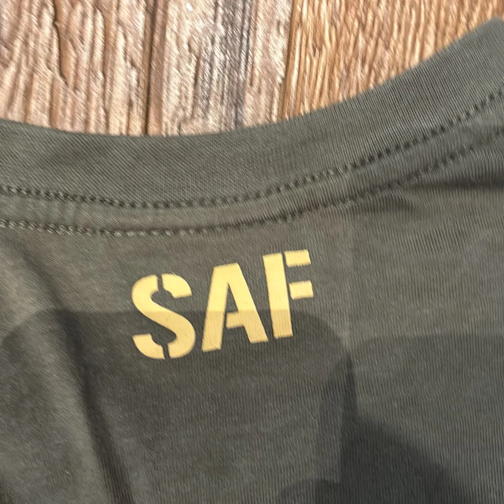 Säljer en olivgrön t-shirt från SAF med texten 'Swedish Armed Forces' i gult på framsidan. T-shirten har korta ärmar och är i klassisk arméstil. Perfekt för vardagsbruk eller som en del av en militärinspirerad outfit. Storlek 160, vilket passar bra för ungdomar eller mindre vuxna.. T-shirts.