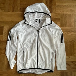 Vit vindjacka från Adidas - Säljer en snygg vit vindjacka/tränings jacka från Adidas i storlek S/M. Jackan har en huva och svarta detaljer som dragkedja och ränder på ärmarna. I mycket bra skick. Passar perfekt både till träning som springning och vardags.