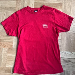 Röd Stüssy t-shirt med draktryck - Säljer en unik röd Stüssy t-shirt i storlek S. Den har ett stort, detaljerat draktryck på ryggen och Stüssy-loggan på framsidan. Perfekt för en avslappnad stil eller streetwear-look. T-shirten är i bomull och har korta ärmar. Köpt på NK, Paul&friends🐉 Kvitto finns