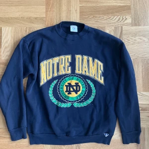 Blå Notre Dame sweatshirt - Säljer en cool blå sweatshirt med Notre Dame-tryck i gult och grönt i heavy weight martial. Tröjan är i bra skick och perfekt för höst och vinter. 