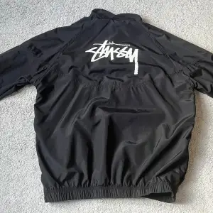 Säljer en svart jacka i mycket bra skick från Stüssy och Nike. Den har en dragkedja framtill och ett stort Stüssy-tryck på ryggen. Perfekt för höst och vår med sin stilrena design och bekväma passform. Passar både till vardags och fest!