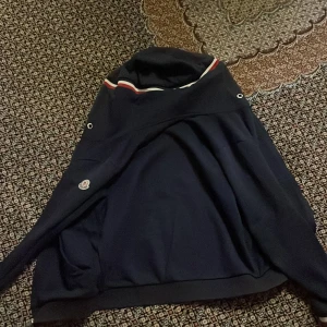 Svart jacka med huva från Moncler - Säljer en snygg svart jacka från Moncler med huva och dragkedja. Den har Moncler-loggan på ärmen och ränder i rött, vitt och blått vid kragen. Perfekt för höst och vår, och den är i mycket bra skick. Passar både till vardags och lite finare tillfällen.   Man kan inte knäppa den så den funkar bara med väst och jag har visat er bild hur de ser ut med väst.