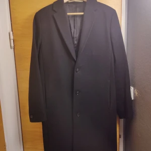 Svart ullrock från Whyred - Säljer en stilren svart ullrock från Whyred i storlek 46 av modell Noren. Perfekt för höst och vinter med sin klassiska design. Tre knappar och dolda fickor fram. Använd men i fint skick!